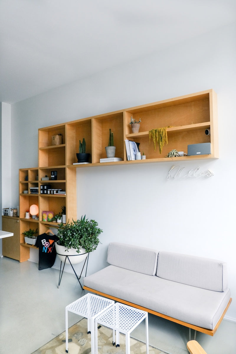 Modern living room with grey sofa against a white wall, modular wood bookshelf rising diagonally from floor left to upper right, leaving a large open wall space above the sofa ideal for wall tile installation