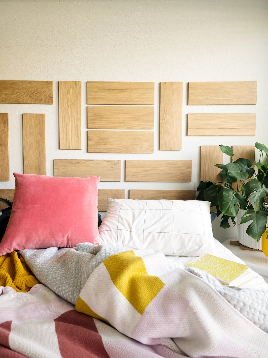 TALAYA Solid Ground Honey Oak tiles installed in a mixed orientation grid above a bed with grey and white bedding, red pillow, colorful striped blanket, and plants in soft natural lighting