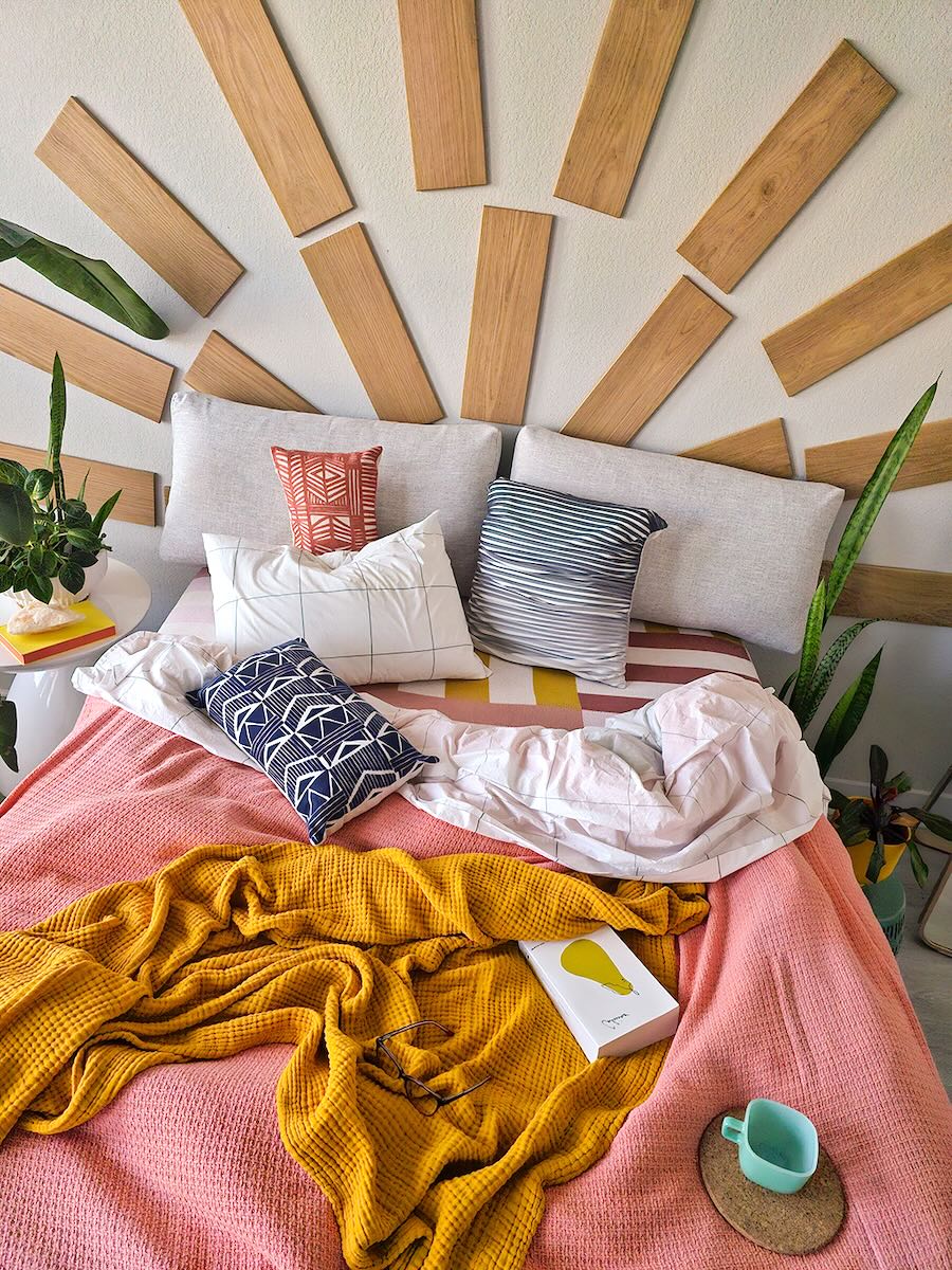 TALAYA Solid Ground tiles in Honey Oak finish arranged in concentric semicircles above a bed, creating a sunburst wall installation in a bright boho bedroom with colorful bedding, green plants, and natural light