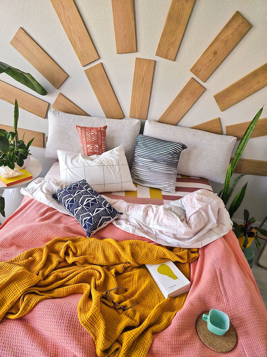 TALAYA Solid Ground tiles in Honey Oak finish arranged in concentric semicircles above a bed, creating a sunburst wall installation in a bright boho bedroom with colorful bedding, green plants, and natural light