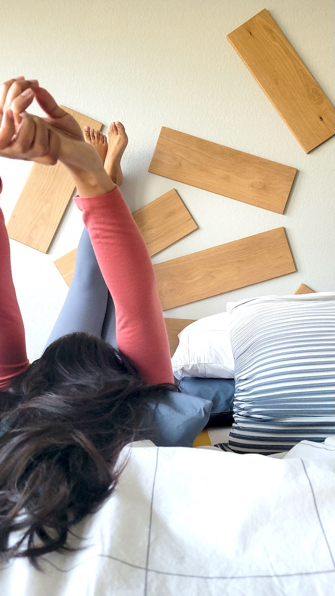 Person lying on a bed with legs up the wall  and arms stretched overhead in celebration, TALAYA Solid Ground Honey Oak tiles installed on wall in two intersecting semicircle patterns
